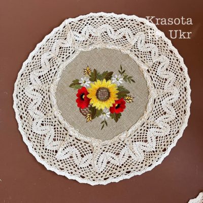 Кругла серветочка з мереживними краями, оздоблена вишивкою яскравого соняшника та червоних маків на світлій тканині.